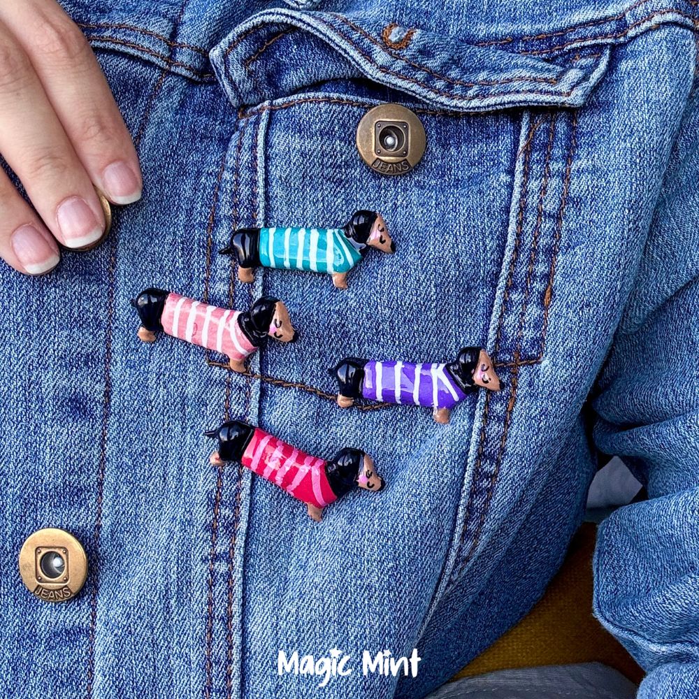 Four dachshund-shaped  air dry clay pins with colorful striped sweaters are pinned onto a blue denim jacket. A hand is visible on the left side of the frame, and the brand name 'Magic Mint' is printed on the lower right of the jacket.